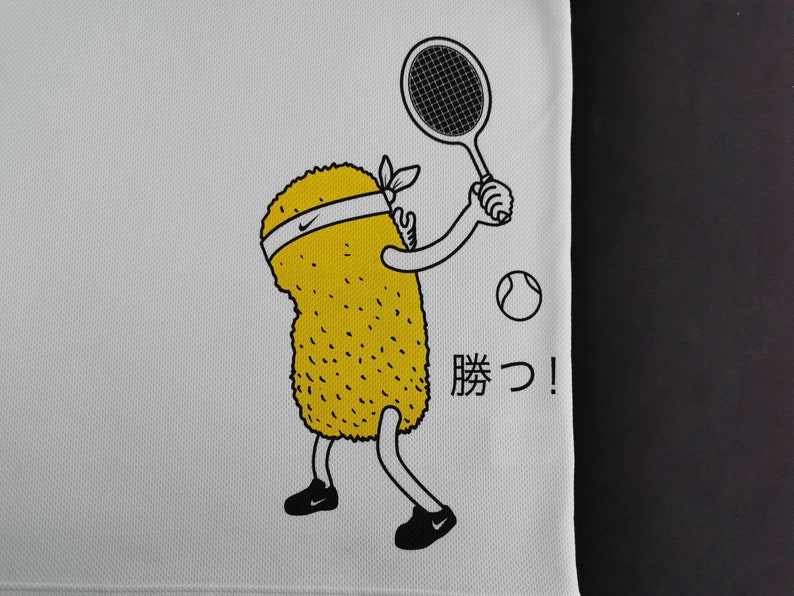 May include: White athletic shirt with a cartoon yellow character playing tennis. The character has a white headband with a Nike logo and black shoes. A tennis ball and racket are also shown. Japanese text "Katsu!" is printed on the shirt.