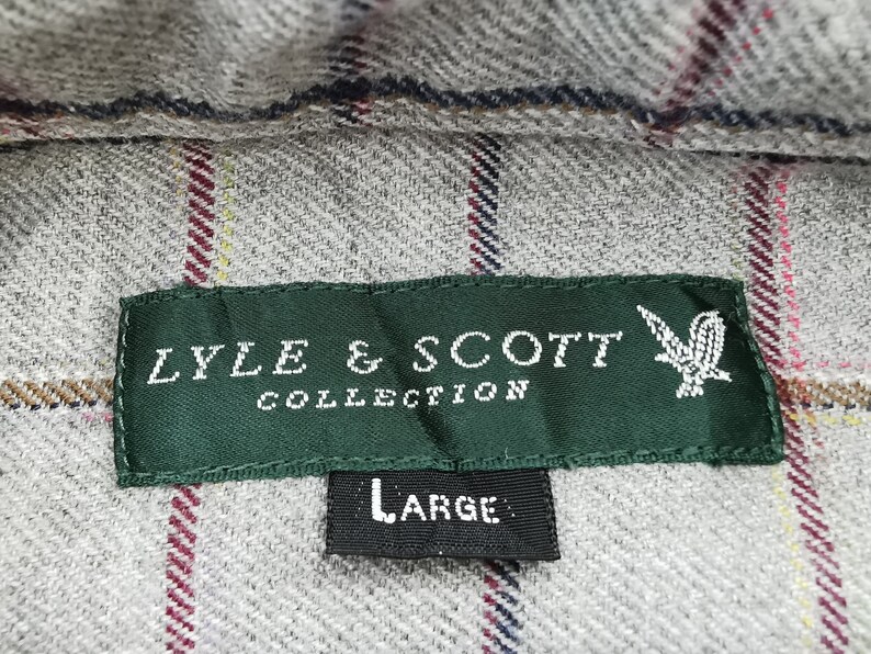 以下が含まれることがあります： グレーのチェック柄の生地のクローズアップ。緑色のラベルには「LYLE & SCOTT COLLECTION」と書かれています。ラベルの下には「LARGE」と書かれた黒いタグがあります。生地には赤、黄、青のストライプがあります。