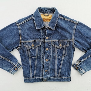 May include: A blue denim jacket with a collared neckline, button-down front, and two chest pockets. The jacket has long sleeves with buttoned cuffs and a gold-colored label at the back of the collar. A classic outerwear garment.