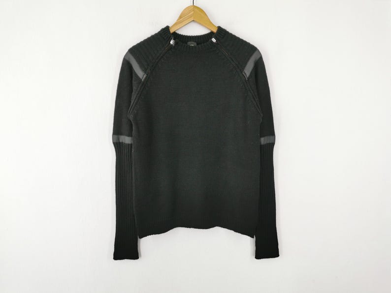 May include: Black sweater with leather-like accents on the shoulders and sleeves. The long-sleeved sweater is hanging on a wooden hanger against a white background. The sweater has a ribbed knit cuff and hem.