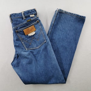 May include: Blue denim jeans with a classic five-pocket design. The jeans feature a straight leg cut and a brown leather patch on the back pocket with the "Wrangler" logo. The jeans are folded and laid flat.