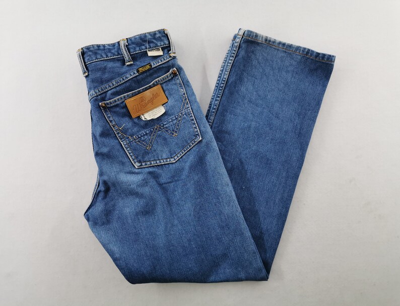 May include: Blue denim jeans with a classic five-pocket design. The jeans feature a straight leg cut and a brown leather patch on the back pocket with the "Wrangler" logo. The jeans are folded and laid flat.