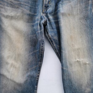 May include: Close-up of blue denim jeans with faded areas and a distressed look. The jeans have a worn appearance with a small tear on the right leg. The denim has a classic five-pocket design.