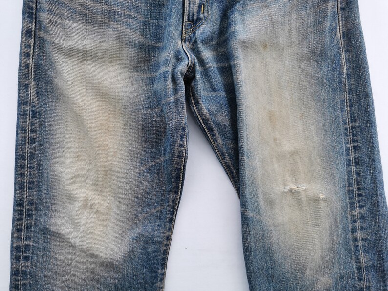 May include: Close-up of blue denim jeans with faded areas and a distressed look. The jeans have a worn appearance with a small tear on the right leg. The denim has a classic five-pocket design.