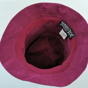 K&ouml;nnte beinhalten: Burgunderfarbener Fischerhut mit strukturiertem Stoff, von innen betrachtet. Die Innenseite hat ein dunkleres Burgunderband und ein Etikett mit dem Markennamen "Kangol" und einem Union-Jack-Logo.
