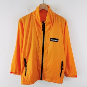 May include: Bright orange jacket with a black zipper and the "Wild Things" logo on the chest. The jacket has a high collar, two side pockets, and black accents. The jacket is hanging on a wooden hanger against a white background.