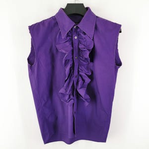 Puede incluir: Una camisa morada sin mangas con la parte delantera con volantes. La camisa tiene un cuello puntiagudo y cierre de botones. La prenda está colgada de una percha negra sobre un fondo blanco. La camisa es de tela lisa.