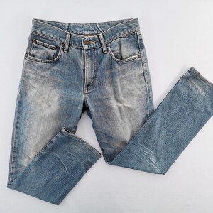 May include: Light blue denim jeans with a faded wash and classic five-pocket design. The jeans have a button and zipper closure, belt loops, and a straight leg cut. The fabric shows signs of wear, adding to the vintage aesthetic.