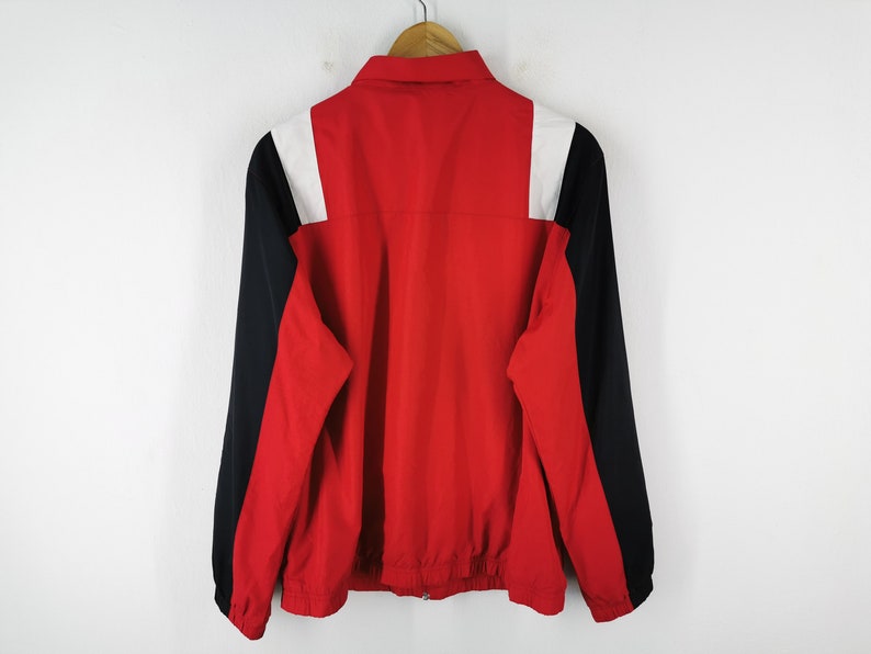 May include: A red, black, and white jacket with a collared neckline and a full-length zipper. The jacket has black sleeves and white shoulder accents. The jacket is hanging on a wooden hanger against a white background.