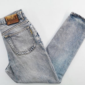 Op de afbeelding: Lichtblauwe jeans met een acid wash en een klassiek ontwerp met vijf zakken. De jeans hebben een bruine leren patch met de merknaam "EDWIN" op de achterkant. De jeans zijn opgevouwen en plat op een witte ondergrond gelegd.