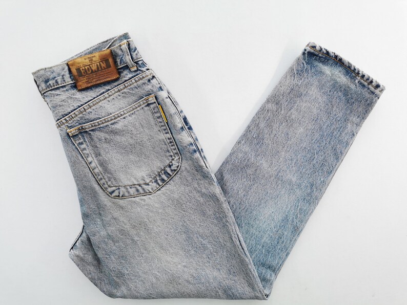 Op de afbeelding: Lichtblauwe jeans met een acid wash en een klassiek ontwerp met vijf zakken. De jeans hebben een bruine leren patch met de merknaam "EDWIN" op de achterkant. De jeans zijn opgevouwen en plat op een witte ondergrond gelegd.