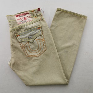 May include: Khaki-colored denim jeans with orange stitching and a True Religion brand label. The jeans feature a classic five-pocket design, with a distinctive horseshoe-shaped stitch on the back pockets. The jeans are folded and laid flat.