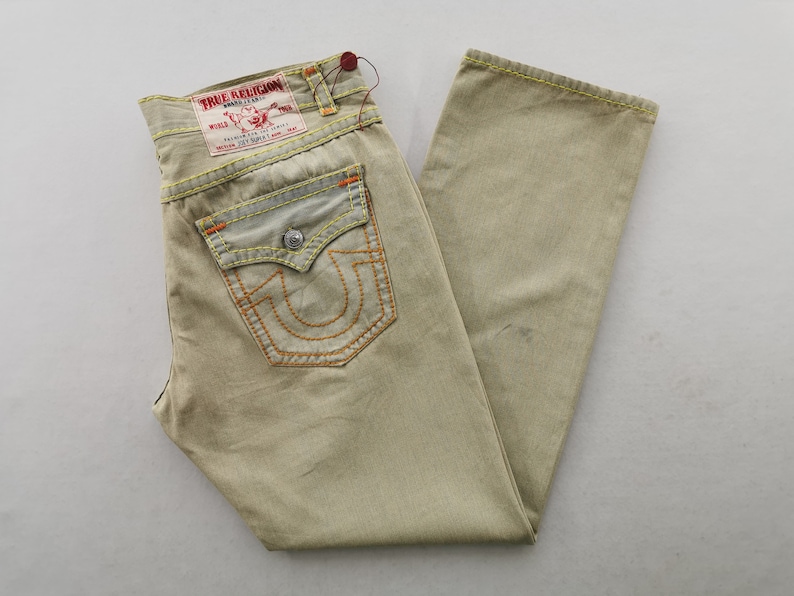 May include: Khaki-colored denim jeans with orange stitching and a True Religion brand label. The jeans feature a classic five-pocket design, with a distinctive horseshoe-shaped stitch on the back pockets. The jeans are folded and laid flat.
