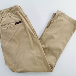 May include: Khaki-colored pants with an elastic waistband and a small back pocket. The pants have a relaxed fit and a small black and red logo on the back. The pants are made of a lightweight material.