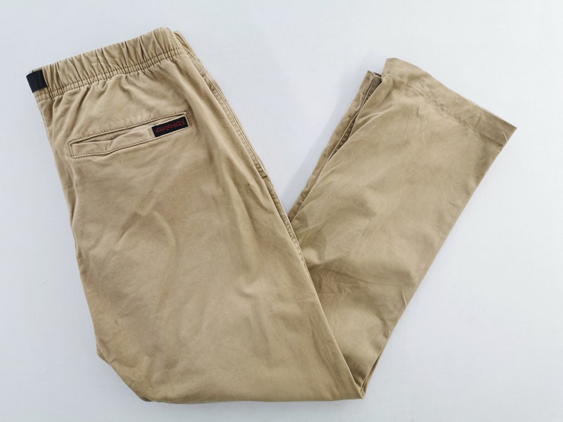 May include: Khaki-colored pants with an elastic waistband and a small back pocket. The pants have a relaxed fit and a small black and red logo on the back. The pants are made of a lightweight material.