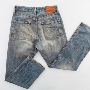 May include: Faded blue denim jeans with a classic five-pocket design. The jeans have a worn appearance with subtle fading and distressing. A brown leather patch is visible on the back waistband. The jeans are laid flat on a white surface.
