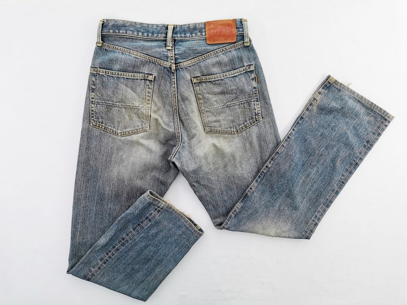 May include: Faded blue denim jeans with a classic five-pocket design. The jeans have a worn appearance with subtle fading and distressing. A brown leather patch is visible on the back waistband. The jeans are laid flat on a white surface.