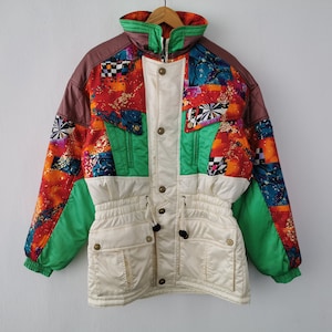 May include: A colorful vintage ski jacket with a high collar and a mix of patterns. The jacket features a cream-colored front with brown, green, and orange accents. It has a drawstring waist and multiple pockets, perfect for winter sports.