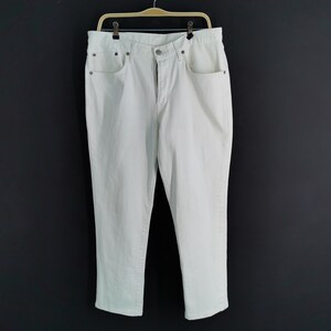May include: Off-white denim jeans hanging on a wooden hanger. The jeans have a classic five-pocket design with a button and zip closure. The trousers are a light colour and appear to be made of a sturdy material.