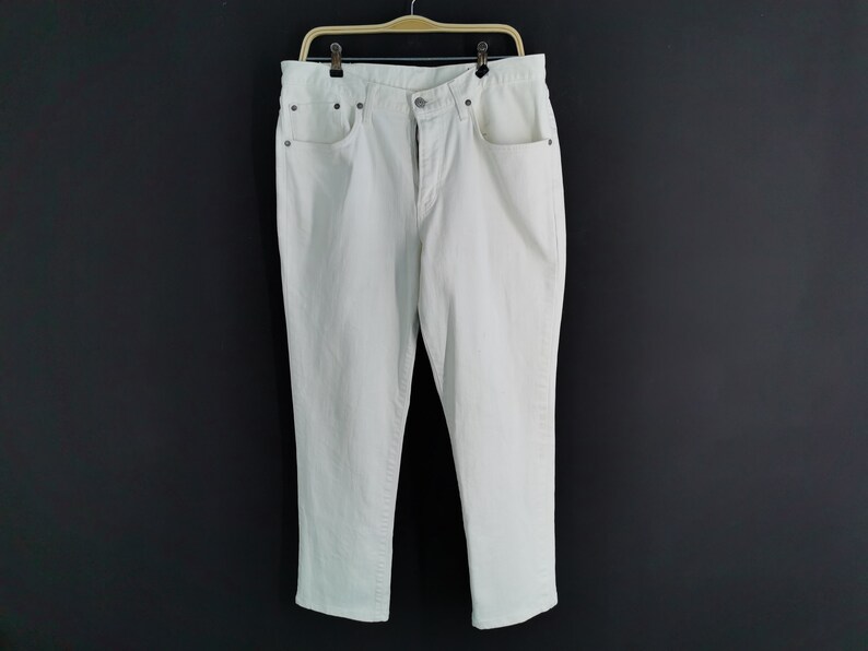 May include: Off-white denim jeans hanging on a wooden hanger. The jeans have a classic five-pocket design with a button and zip closure. The trousers are a light colour and appear to be made of a sturdy material.