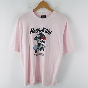 May include: A light pink short-sleeve t-shirt featuring a graphic of Hello Kitty wearing a black cap and sunglasses while on a skateboard. The text reads Hello Kitty, Have fun!, and TODAY, TOMORROW AND ALWAYS. The shirt is displayed on a wooden hanger.
