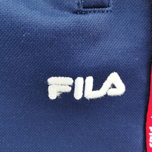 Puede incluir: Primer plano de ropa deportiva azul marino con el nombre de la marca "FILA" bordado en blanco. Una franja roja con el nombre de la marca en blanco recorre verticalmente el lateral.