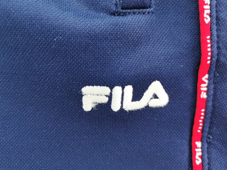 Puede incluir: Primer plano de ropa deportiva azul marino con el nombre de la marca "FILA" bordado en blanco. Una franja roja con el nombre de la marca en blanco recorre verticalmente el lateral.