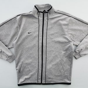 Puede incluir: Chaqueta deportiva gris con cremallera, ribete negro y un peque&ntilde;o logotipo de Nike en el pecho. La chaqueta tiene cuello alto y dos bolsillos delanteros. Las mangas tienen pu&ntilde;os acanalados.