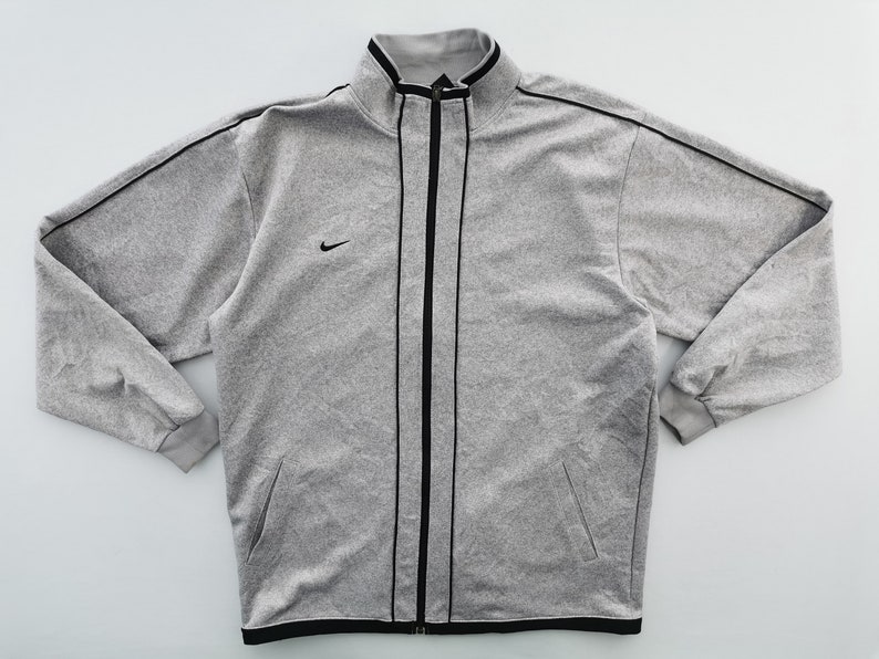 Puede incluir: Chaqueta deportiva gris con cremallera, ribete negro y un peque&ntilde;o logotipo de Nike en el pecho. La chaqueta tiene cuello alto y dos bolsillos delanteros. Las mangas tienen pu&ntilde;os acanalados.