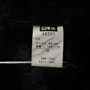 Puede incluir: Primer plano de una etiqueta de ropa Edwin. La etiqueta blanca tiene texto negro, incluyendo el nombre de la marca, detalles de la talla y c&oacute;digos de producto. La etiqueta est&aacute; sobre un fondo negro texturizado.