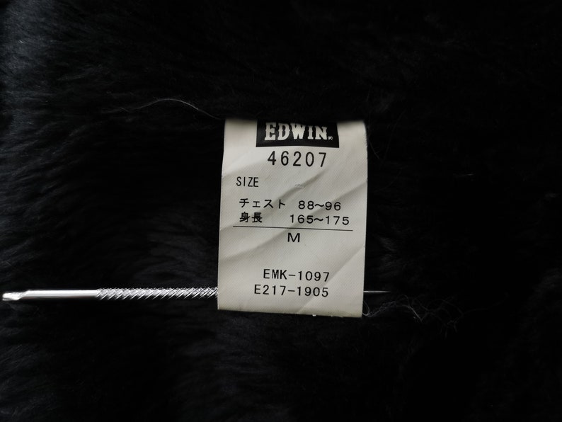 Puede incluir: Primer plano de una etiqueta de ropa Edwin. La etiqueta blanca tiene texto negro, incluyendo el nombre de la marca, detalles de la talla y c&oacute;digos de producto. La etiqueta est&aacute; sobre un fondo negro texturizado.