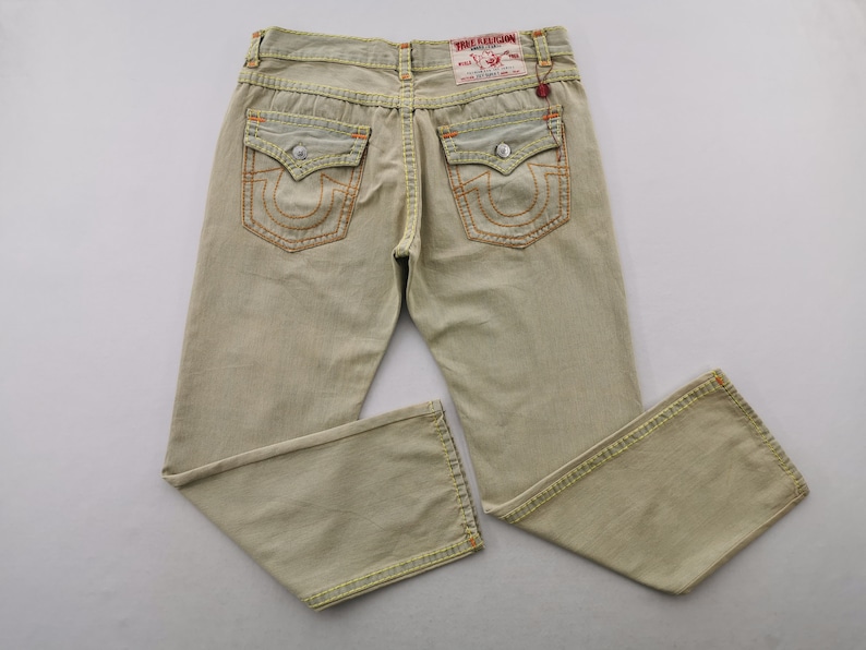 May include: Light tan denim jeans with orange stitching and a classic five-pocket design. Features the True Religion brand label on the back pocket. The jeans have a relaxed fit and are suitable for casual wear.