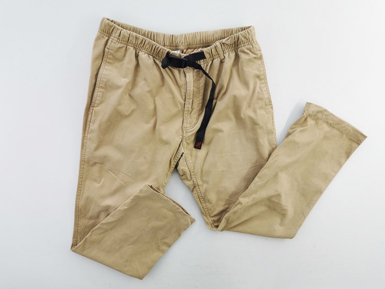 May include: Khaki-colored pants with an elastic waistband and a black belt. The pants have two front pockets and a relaxed fit. The belt has a small red tag. The pants are made of a durable fabric.