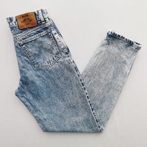 Puede incluir: Vaqueros de mezclilla con lavado &aacute;cido con un dise&ntilde;o cl&aacute;sico de cinco bolsillos. Los vaqueros presentan un lavado azul claro con una apariencia descolorida y texturizada. Un parche de cuero marr&oacute;n es visible en la cintura trasera. Los vaqueros est&aacute;n doblados sobre una superficie blanca.