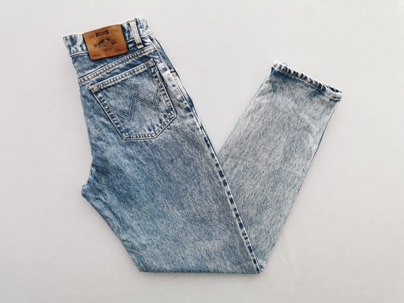 Puede incluir: Vaqueros de mezclilla con lavado &aacute;cido con un dise&ntilde;o cl&aacute;sico de cinco bolsillos. Los vaqueros presentan un lavado azul claro con una apariencia descolorida y texturizada. Un parche de cuero marr&oacute;n es visible en la cintura trasera. Los vaqueros est&aacute;n doblados sobre una superficie blanca.