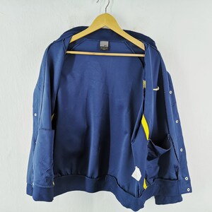 May include: Navy blue zip-up jacket with yellow detailing and a small Nike logo. The jacket features a ribbed collar and cuffs, and two front pockets. It is hanging on a wooden hanger against a white backdrop.