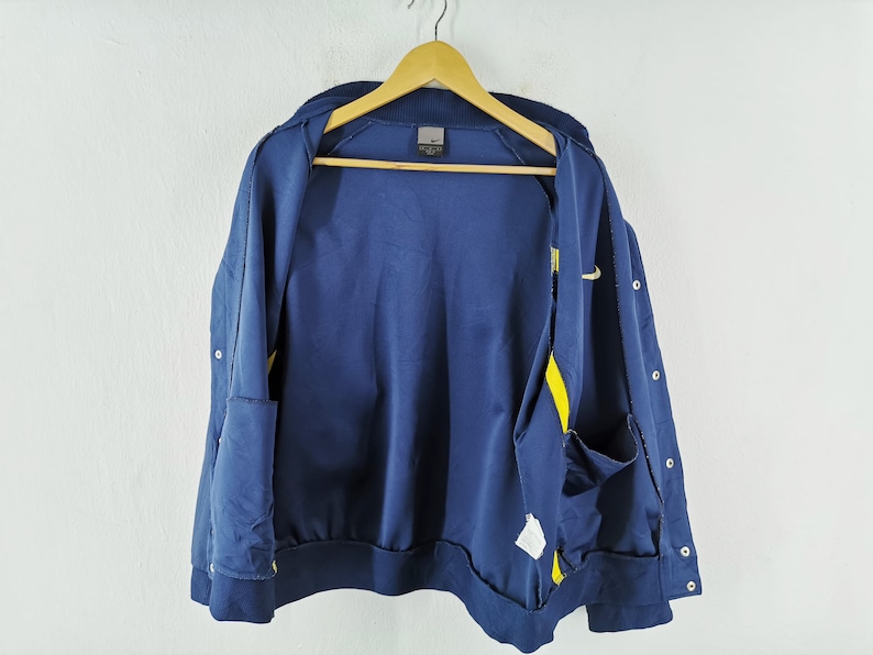 May include: Navy blue zip-up jacket with yellow detailing and a small Nike logo. The jacket features a ribbed collar and cuffs, and two front pockets. It is hanging on a wooden hanger against a white backdrop.