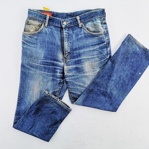 May include: Faded blue denim jeans with a classic five-pocket design. The jeans show signs of wear and tear, with distressed areas and a light wash. The tag is red with white text. The jeans are laid flat on a white surface.