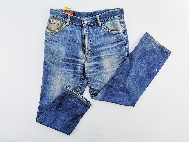May include: Faded blue denim jeans with a classic five-pocket design. The jeans show signs of wear and tear, with distressed areas and a light wash. The tag is red with white text. The jeans are laid flat on a white surface.