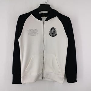 Puede incluir: Sudadera con capucha blanca con cremallera y mangas y capucha negras. Presenta el texto "SINCE 1947 VON DUTCH LOS ANGELES GENUINE VINTAGE" en la parte delantera, junto con un logotipo de escudo. Incluye un bolsillo delantero.