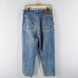 Op de afbeelding: Lichtblauwe spijkerbroek hangend aan een houten hanger. De jeans heeft een klassiek ontwerp met vijf zakken, een leren patch met het "Wrangler"-logo en een rood label op de achterzak. De jeans is vervaagd en heeft een versleten uitstraling.