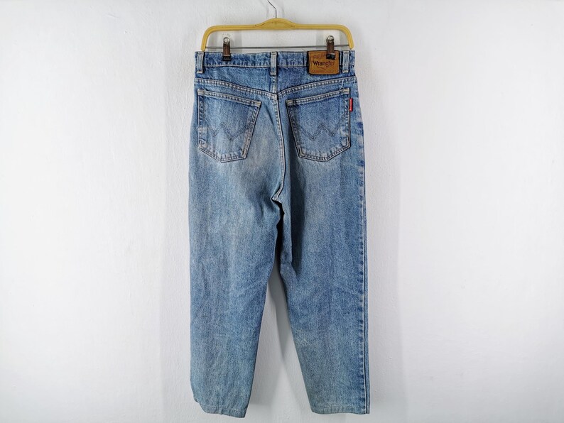 Op de afbeelding: Lichtblauwe spijkerbroek hangend aan een houten hanger. De jeans heeft een klassiek ontwerp met vijf zakken, een leren patch met het "Wrangler"-logo en een rood label op de achterzak. De jeans is vervaagd en heeft een versleten uitstraling.