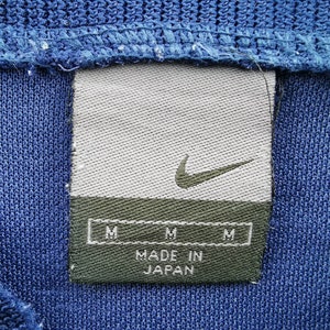 May include: A close-up shot of blue knitwear with a Nike label. The label is grey with a green base, displaying the Nike swoosh and "MADE IN JAPAN". It also features three "M" size indicators.