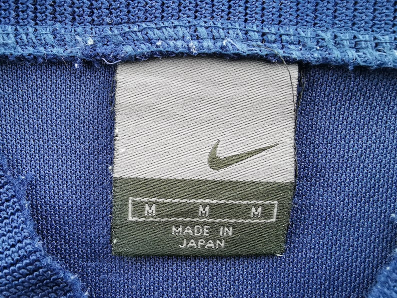 May include: A close-up shot of blue knitwear with a Nike label. The label is grey with a green base, displaying the Nike swoosh and "MADE IN JAPAN". It also features three "M" size indicators.