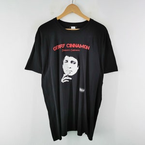 May include: Black t-shirt featuring a graphic of a person and the text "Gerry Cinnamon". The text is in red, and the graphic is in white. The shirt is displayed on a wooden hanger.