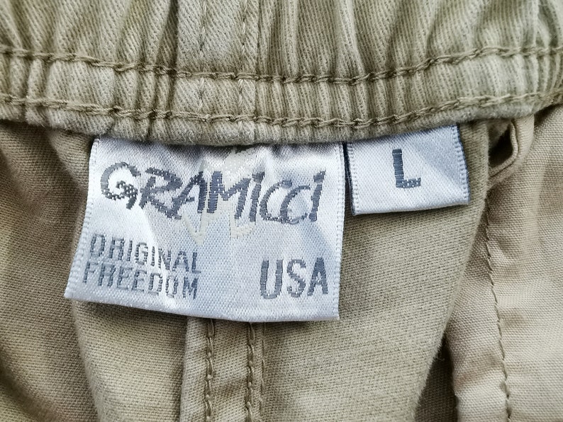 May include: Close-up of a tan fabric label with the brand name "GRAMICCI" and the words "ORIGINAL FREEDOM USA". The label also has a size "L" tag.