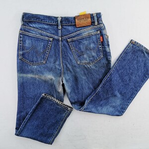 May include: Blue denim jeans with a classic five-pocket design. The jeans feature a faded wash and a leather patch with the brand name "EDWIN". The back pockets have a distinctive "W" stitch. A small red tag is visible.