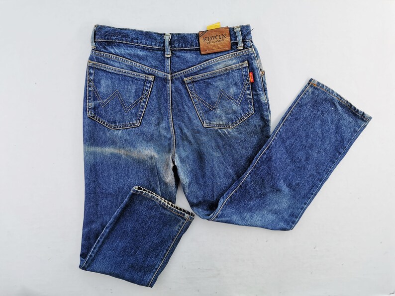 May include: Blue denim jeans with a classic five-pocket design. The jeans feature a faded wash and a leather patch with the brand name "EDWIN". The back pockets have a distinctive "W" stitch. A small red tag is visible.