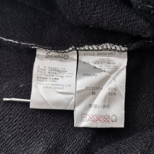 May include: A close-up shot of a black garment with attached labels. The labels show the "Sanrio LICENSE" logo, product details, and fabric content: 65% polyester, 35% cotton. The style number of the garment is B6525KP-2.