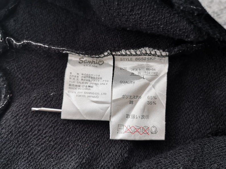 May include: A close-up shot of a black garment with attached labels. The labels show the "Sanrio LICENSE" logo, product details, and fabric content: 65% polyester, 35% cotton. The style number of the garment is B6525KP-2.